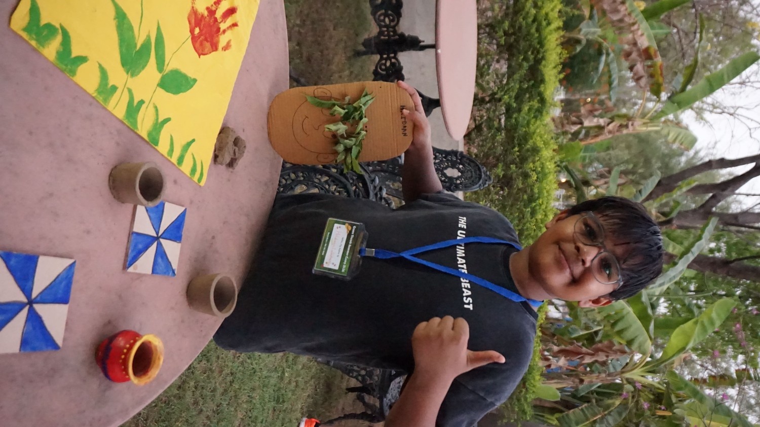 A smiling boy giving a thumbs up while standing behind a table displaying his leaf craft, painted pots, and tiles