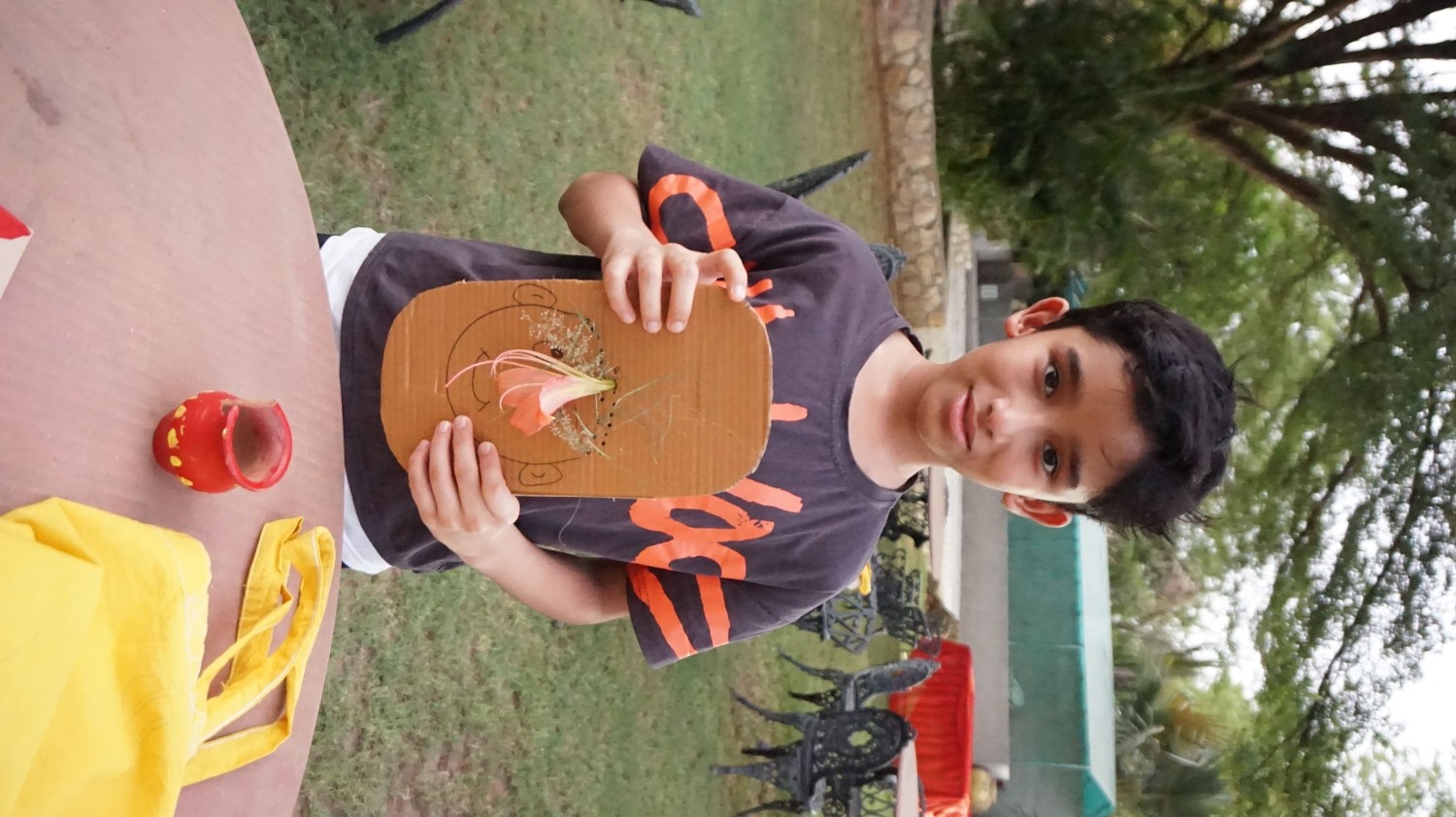 A boy in a black graphic tee holding a cardboard face craft featuring a pink flower placed as the nose