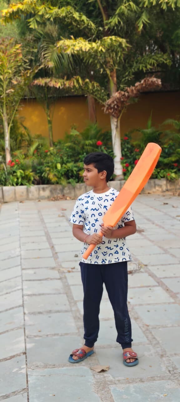 A young boy standing outdoors holding a large orange plastic cricket bat, getting ready to play a game