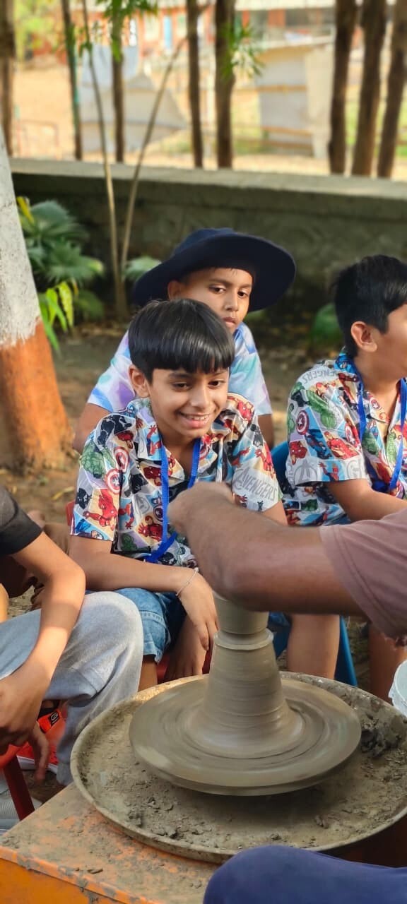 A smiling boy in an Avengers shirt watching an instructor shape clay on a pottery wheel at Tripada School summer camp