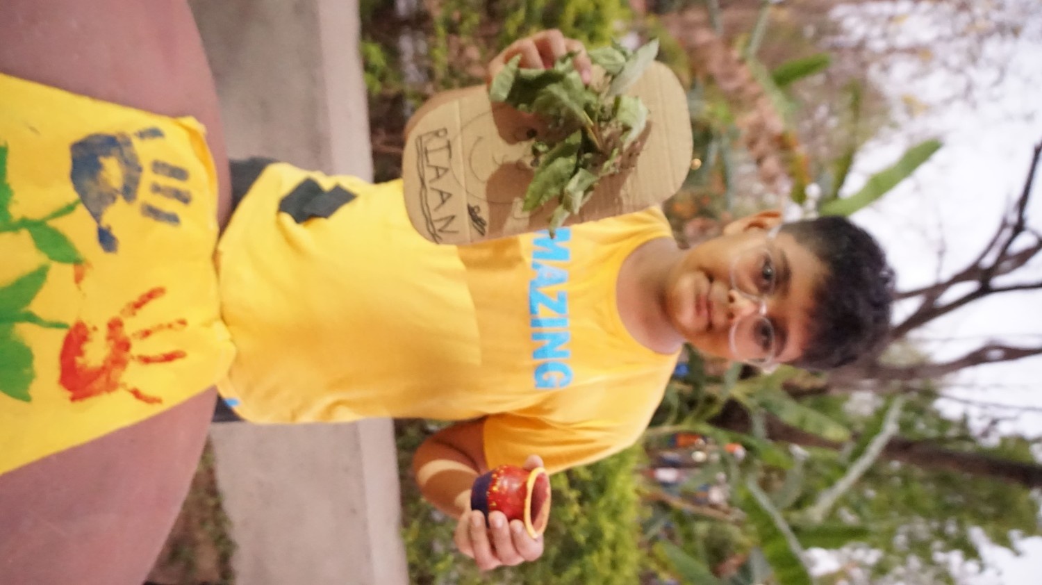 A boy in a yellow shirt named Riaan holding a cardboard face craft with green leaves and a painted clay pot