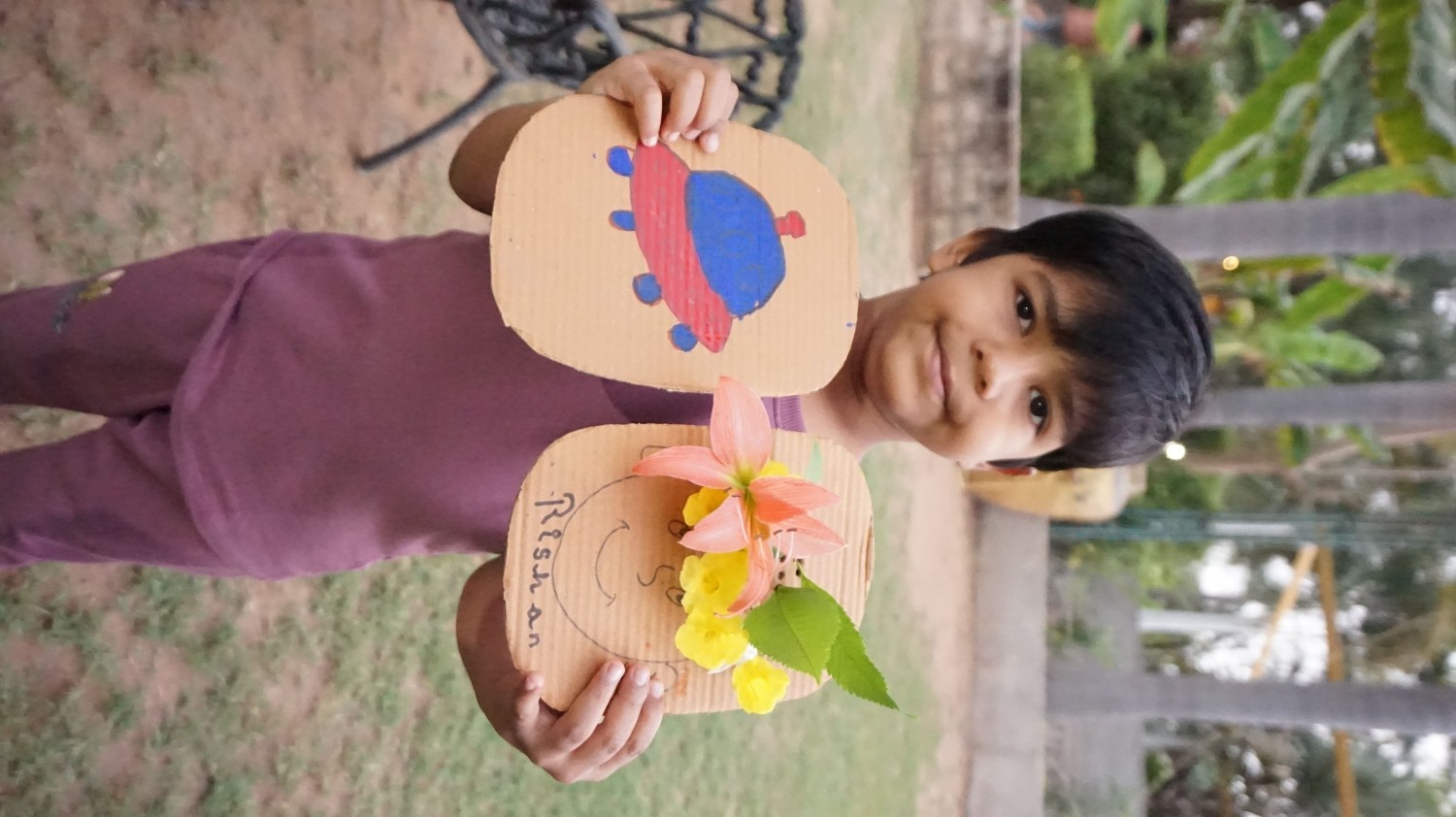 A boy named Rishan holding two cardboard crafts: one painted like a UFO and one decorated with real flowers