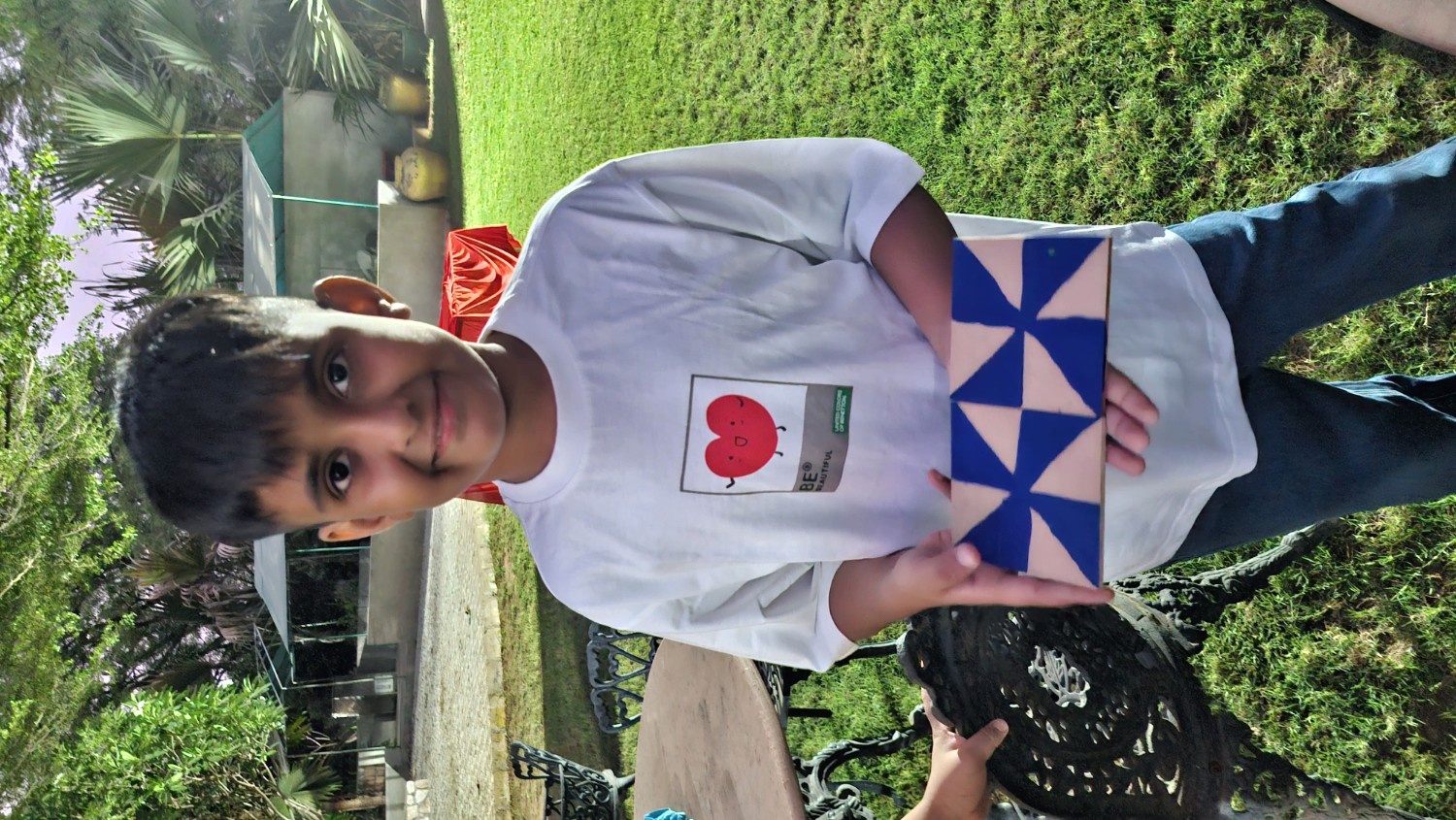 A smiling boy holding up a cardboard tile painted with a blue and white geometric pattern outdoors