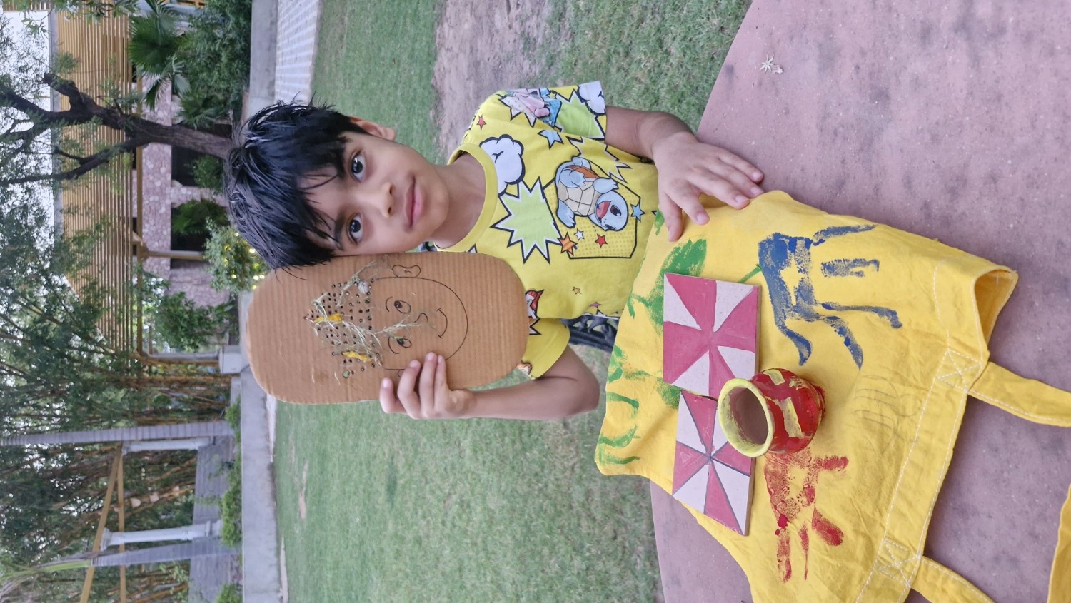 A young boy proudly displaying a cardboard face craft, painted geometric tiles, and a handprint tote bag