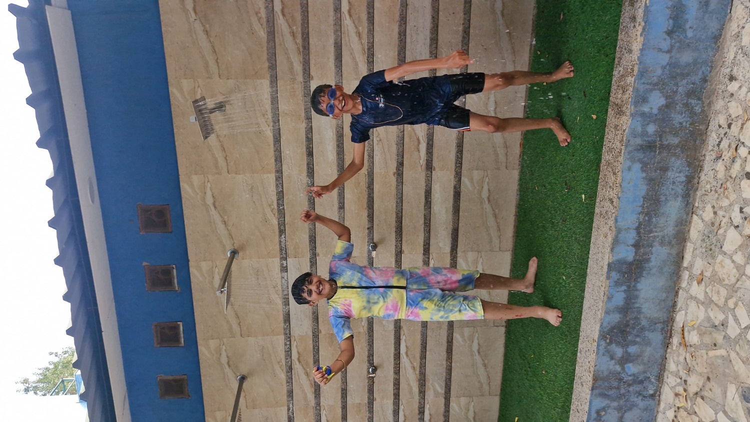 Two smiling boys having fun and cooling off under outdoor rainfall showers by the pool