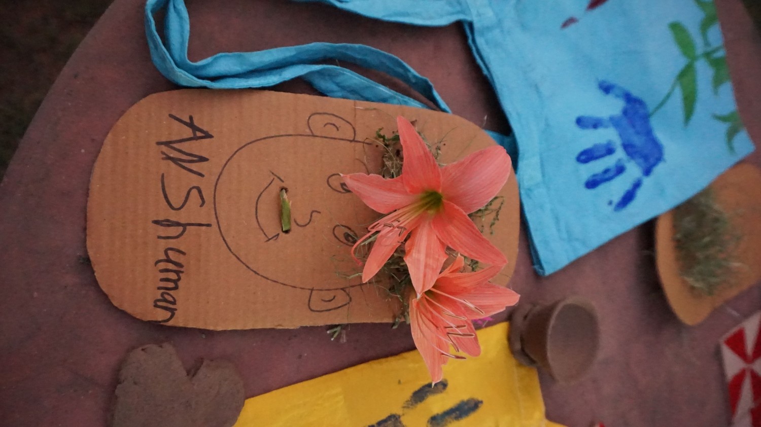 A cardboard face craft with the name Anshuman, decorated with two large pink flowers for hair