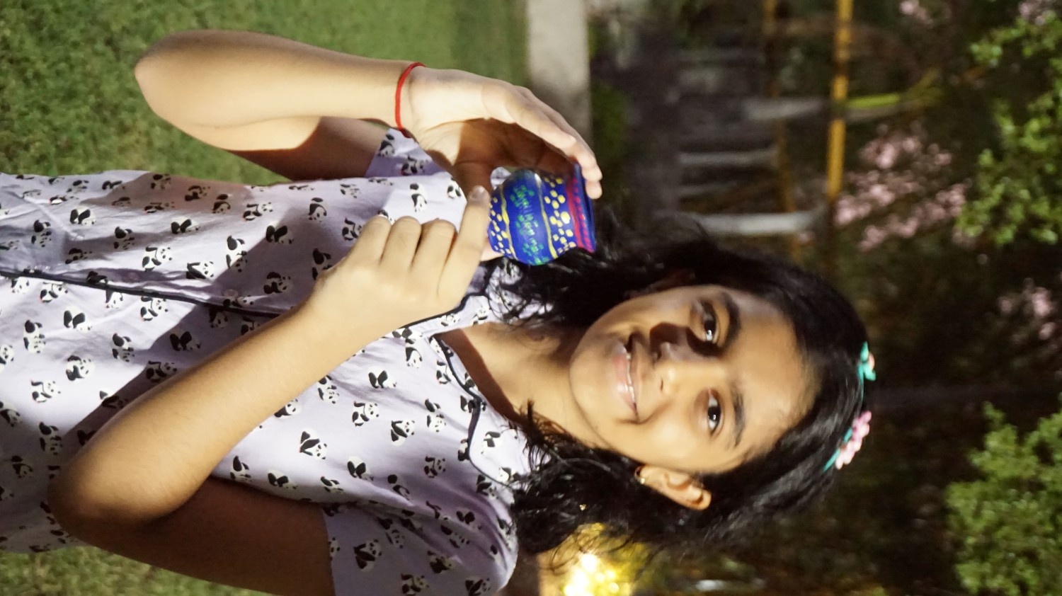 A smiling girl outdoors at night holding up a small clay pot painted dark blue with yellow dots
