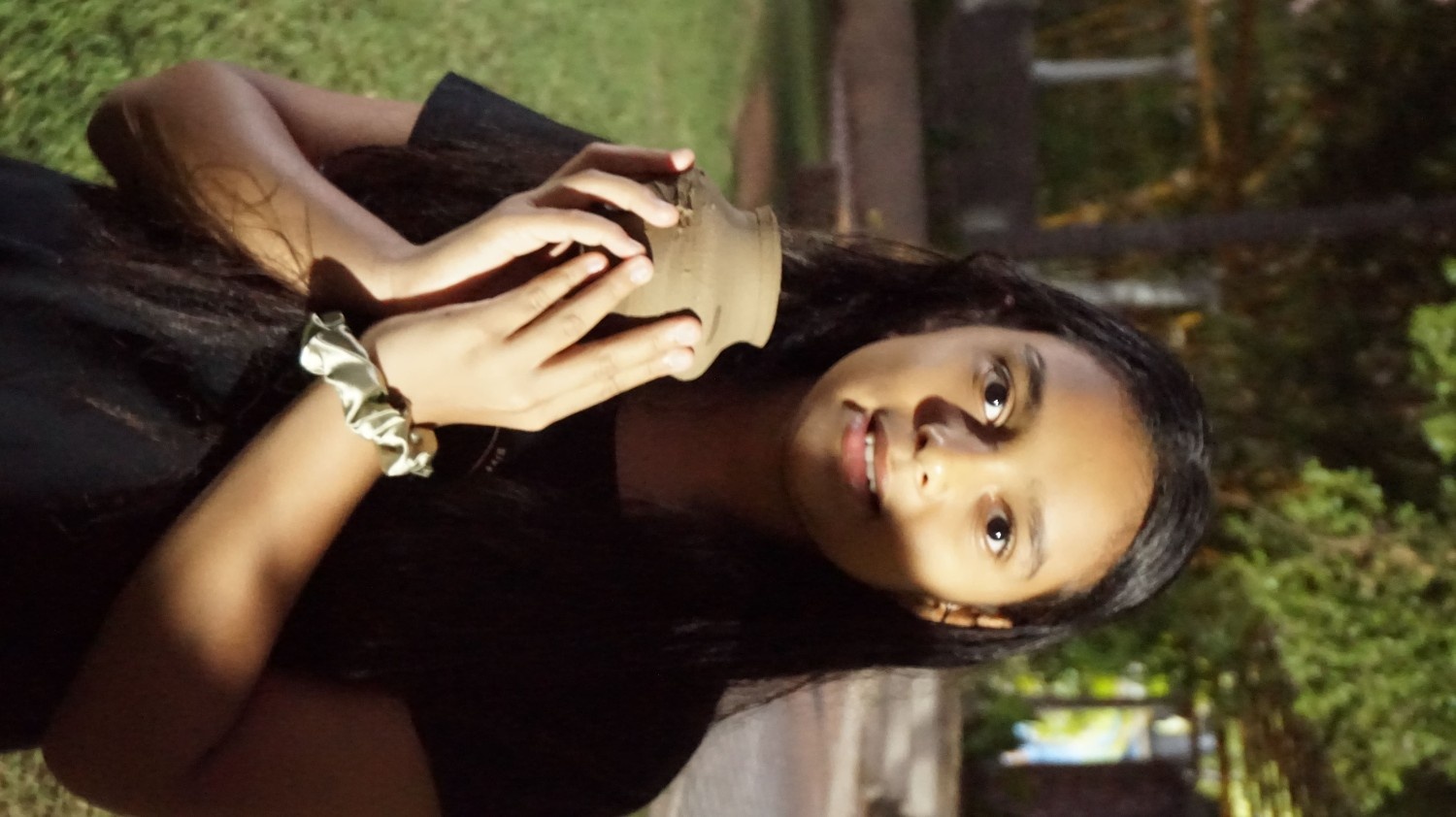 A girl standing outdoors at night, carefully holding a freshly made, unpainted clay pot in her hands