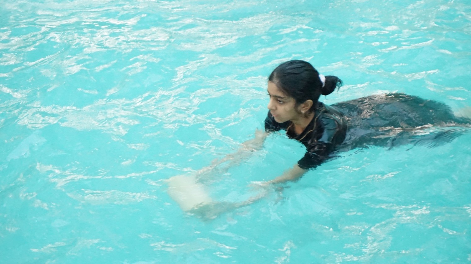 A young girl practicing swimming in an outdoor pool while holding onto a white foam kickboard