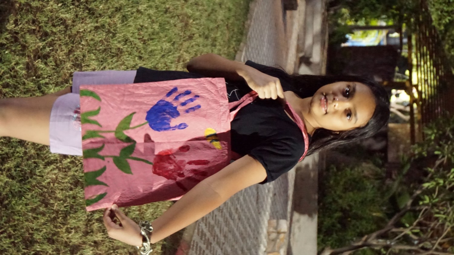 A young girl standing outdoors at night showing off a pink canvas tote bag decorated with blue and red handprint flowers