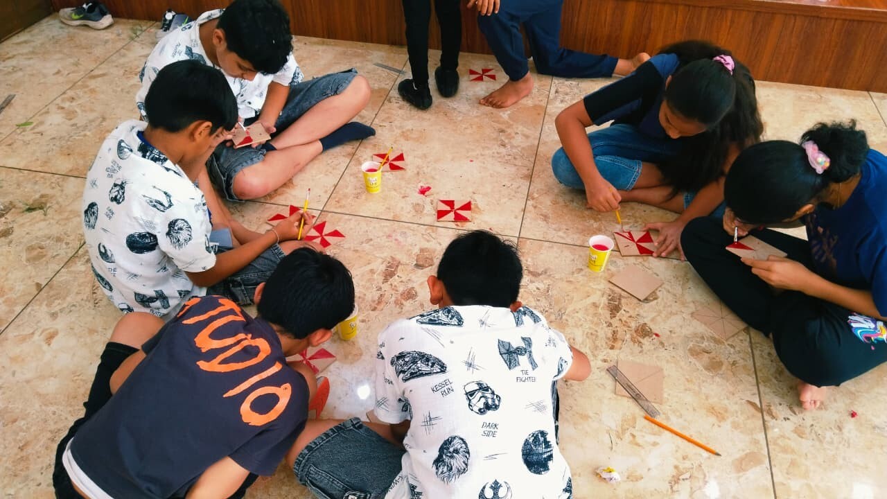 A group of children sitting in a circle painting red and white geometric patterns on square tiles