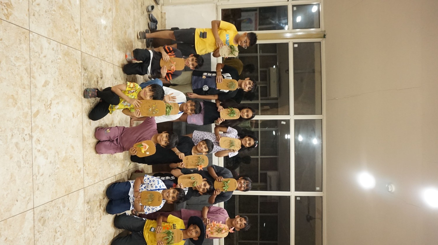 A large group of smiling Tripada School summer campers posing indoors, all holding up their handmade cardboard face crafts