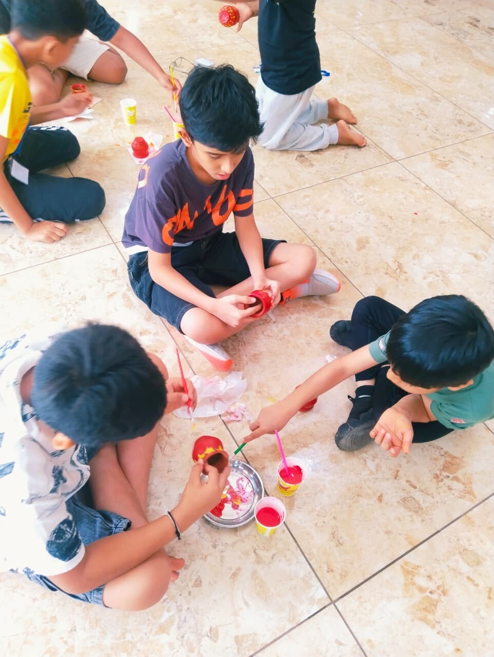 Children sitting on the floor indoors painting small clay pots with red paint