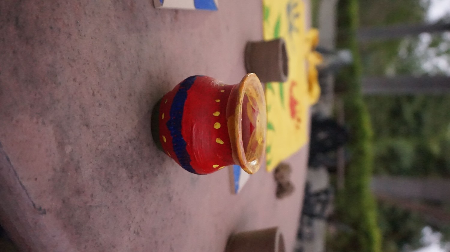 A small clay pot painted bright red with yellow dots and a blue stripe sitting on a table outdoors