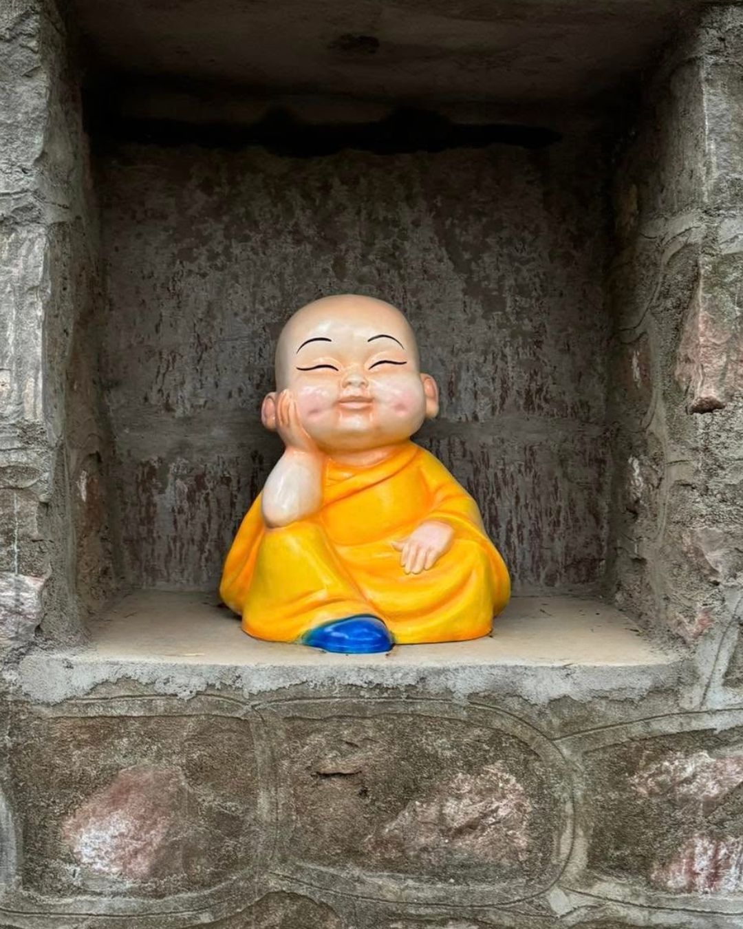 Little monk statue in yellow robe resting cheek on hand in stone wall niche
